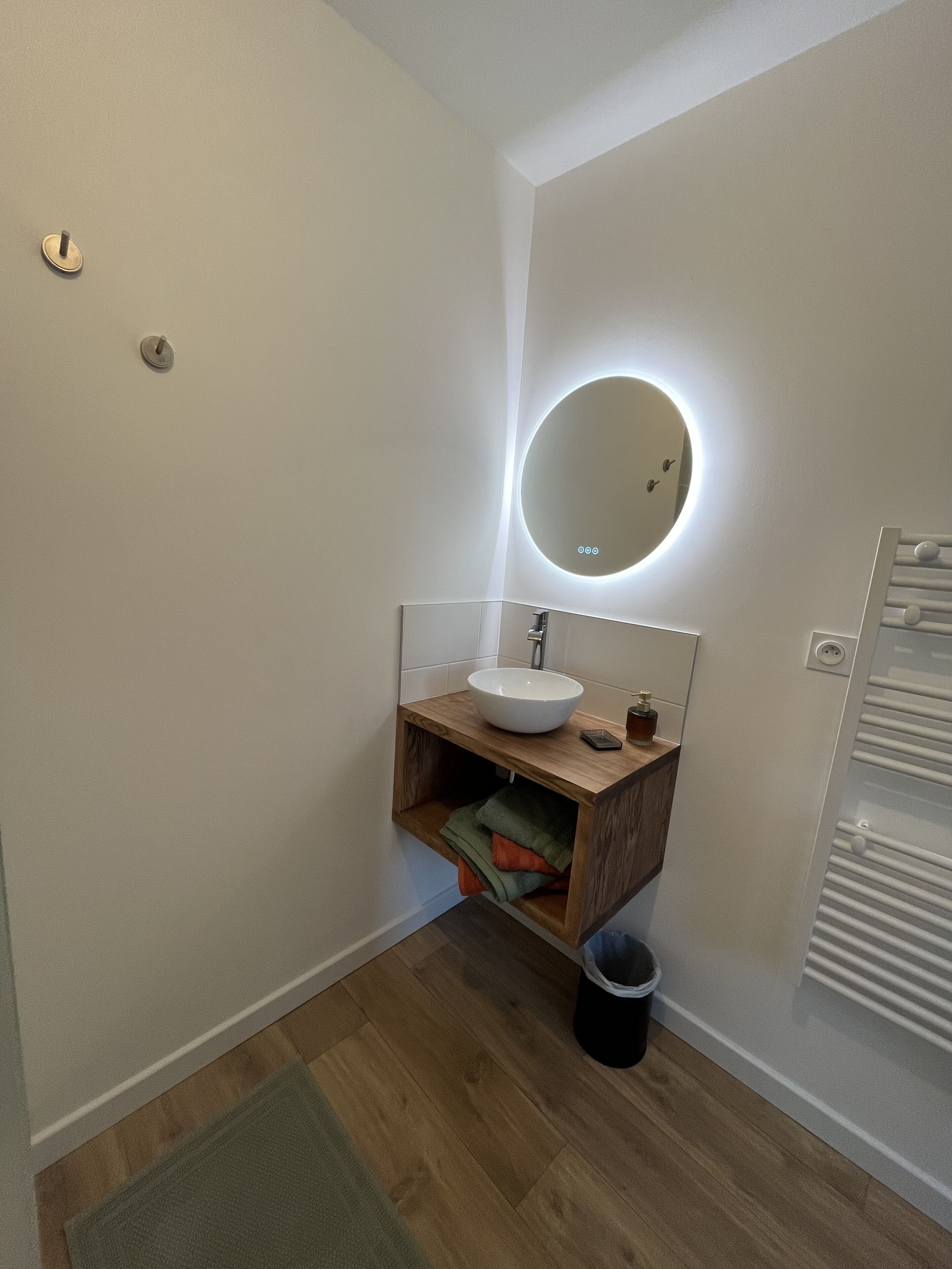 Een moderne badkamer met een houten wastafelmeubel, een ronde spiegel met verlichting, een witte wasbak en een handdoekrek.