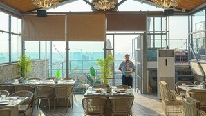 Un comedor interior con mobiliario de mimbre, ventanas amplias y vistas a la ciudad.