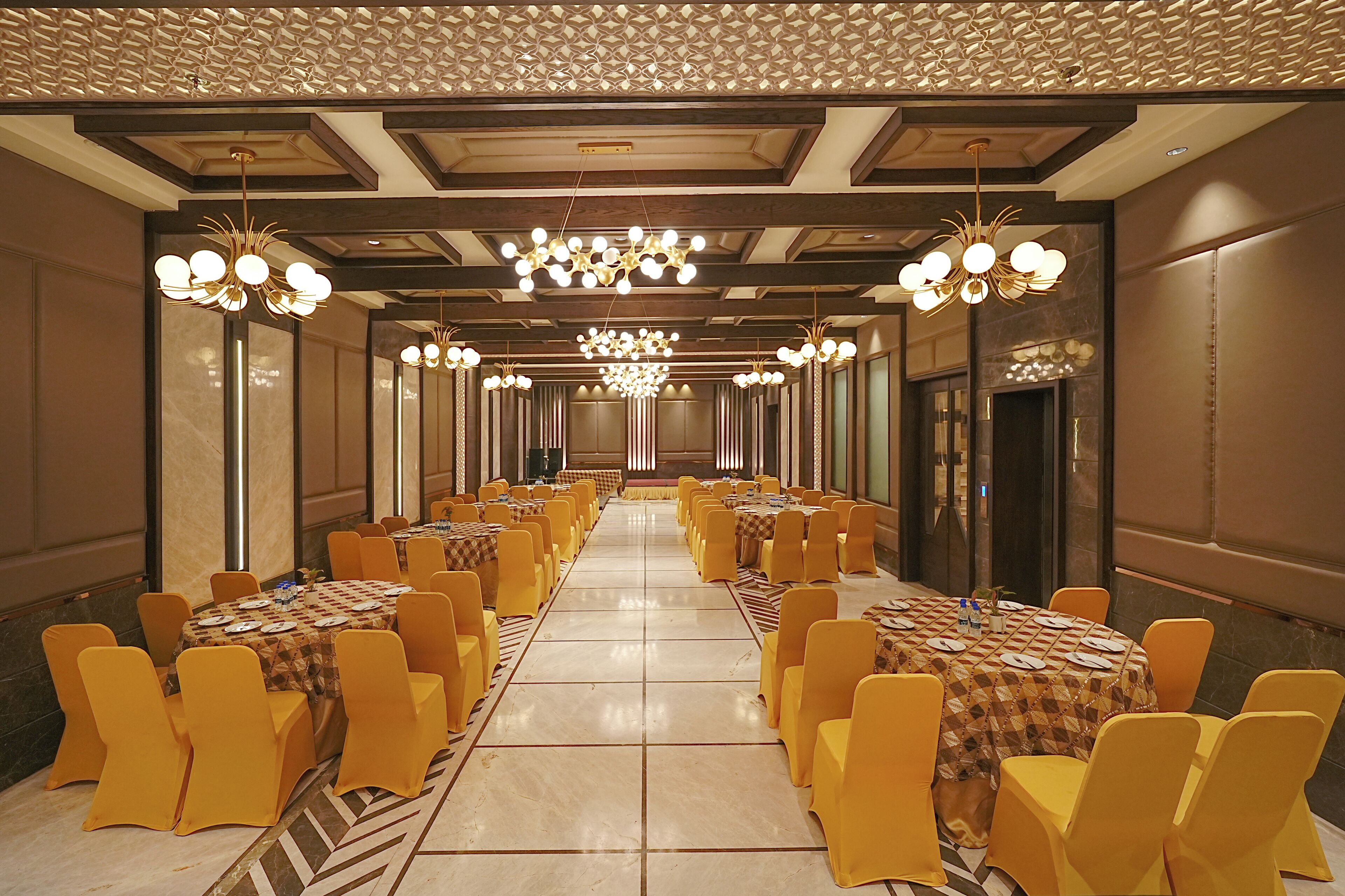 A banquet hall with round tables and yellow chairs, large chandeliers, and a patterned ceiling.