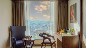 Habitación de hotel con un ventanal grande, un escritorio con una silla, una mesita con frascos y un cuadro en la pared.