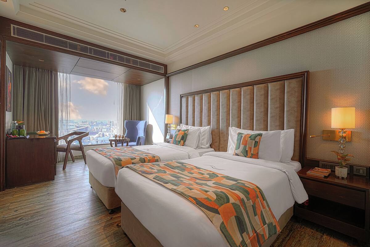 A hotel room with two beds, a desk, a chair, and a large window offering a city view.
