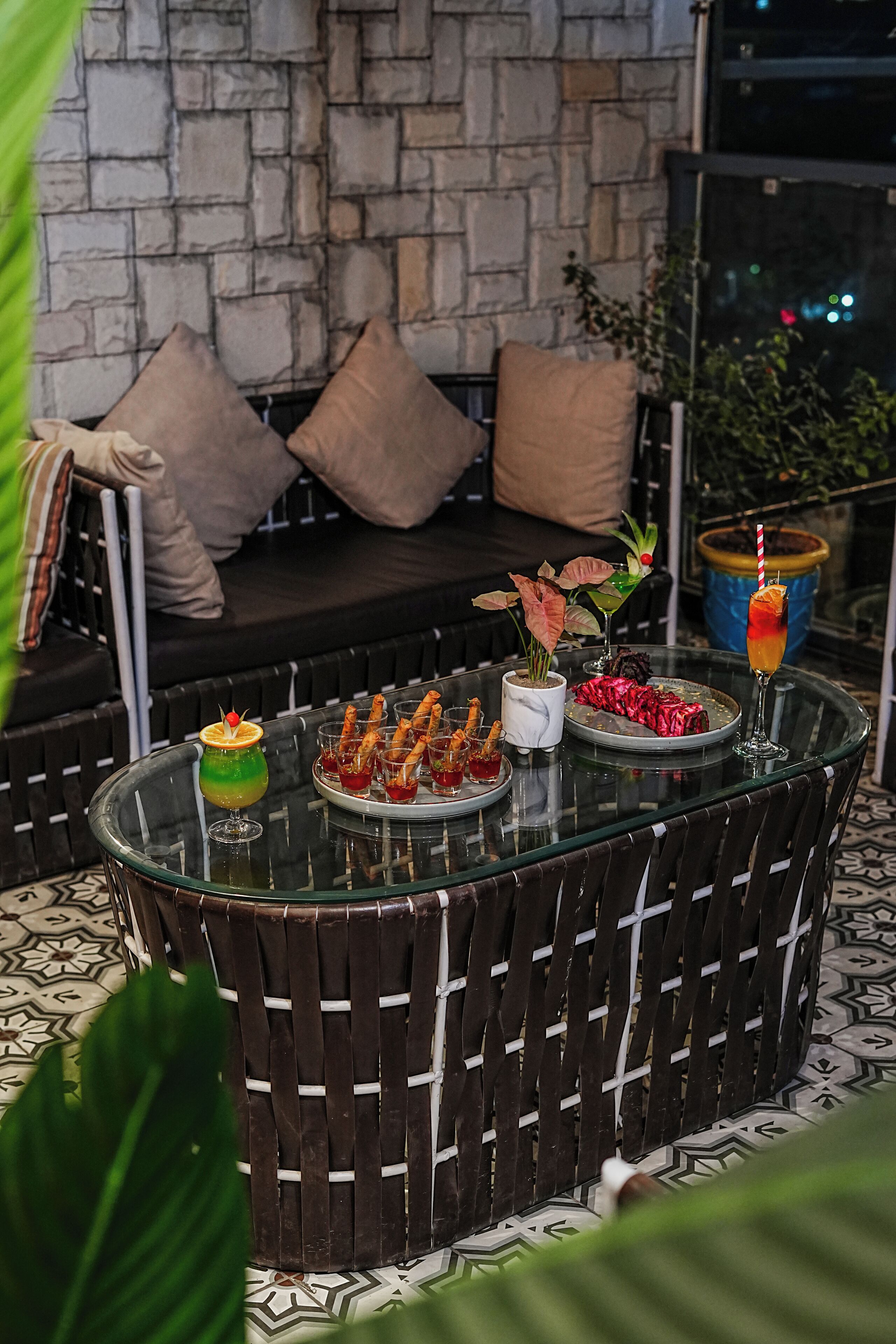 Une table avec un plateau en verre, sur laquelle sont disposés de la nourriture et des boissons, un mur en pierre et un canapé avec des coussins.