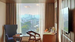 Un cuarto de hotel con un ventanal grande, un sillón azul, una silla de madera, una mesita redonda y un televisor montado en la pared.