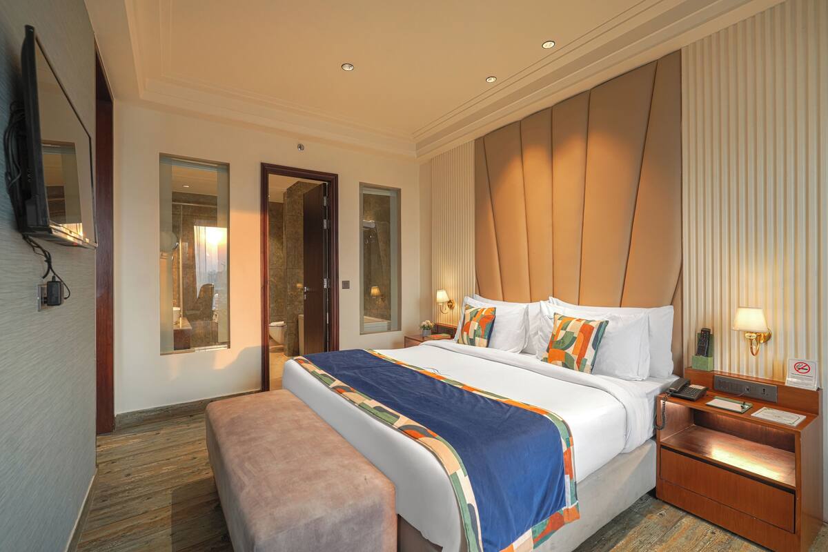 A hotel room with a large bed, a bench, a bedside table with a lamp, and a mirror on the wall.