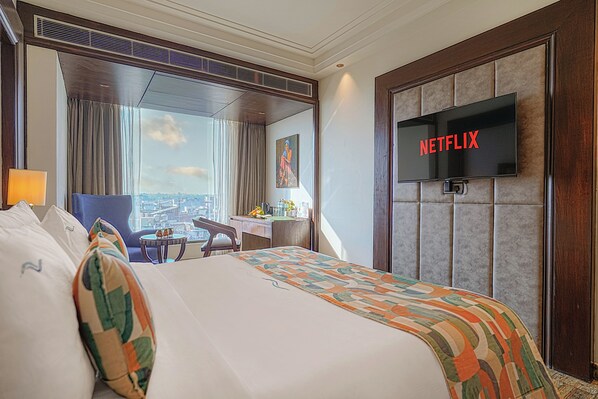 Una habitación de hotel con una cama grande, un escritorio, una silla, un televisor montado en la pared y vistas a la ciudad a través de la ventana.