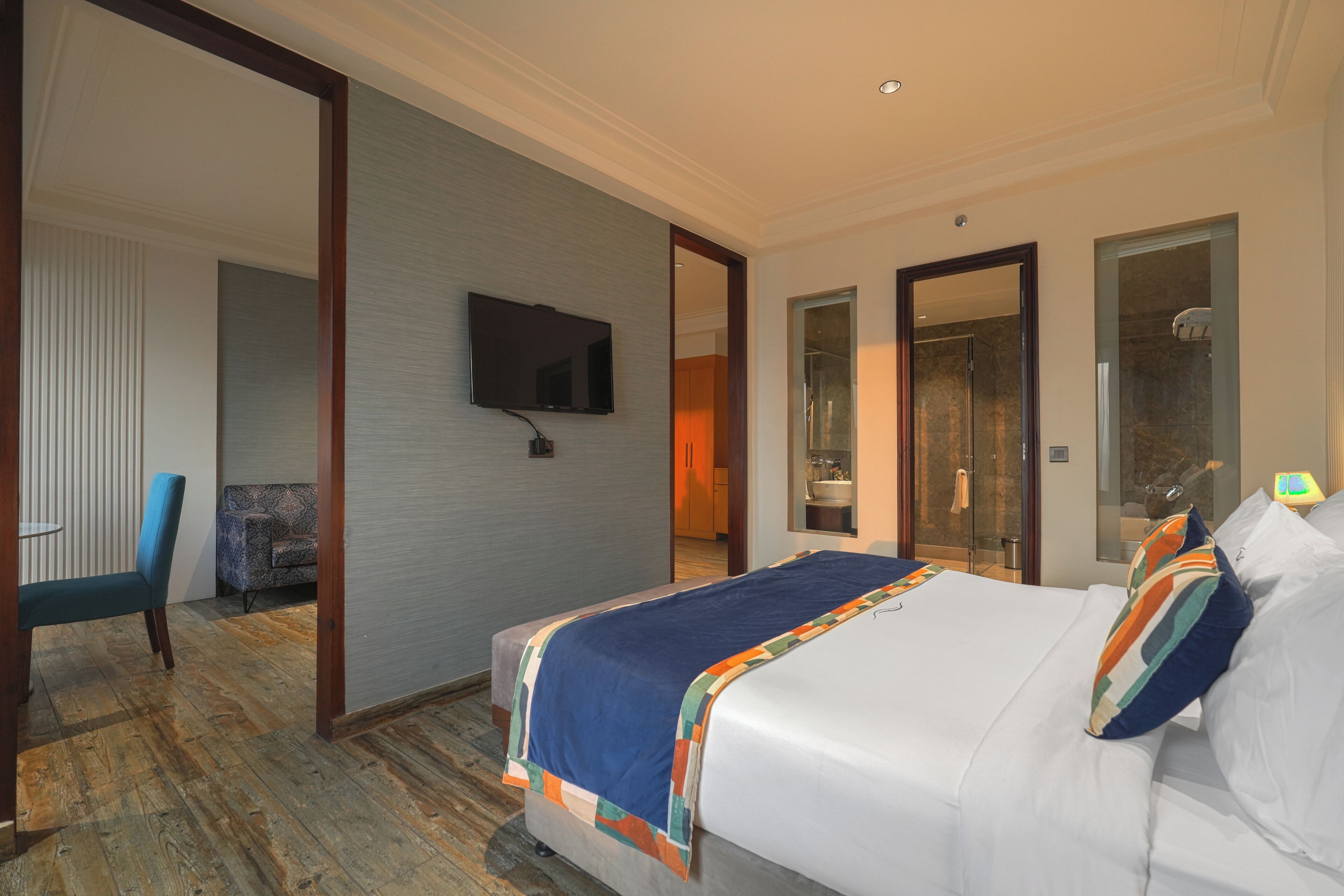 A hotel room with a large bed, a television mounted on the wall, a seating area with a chair, and a bathroom with a glass door.