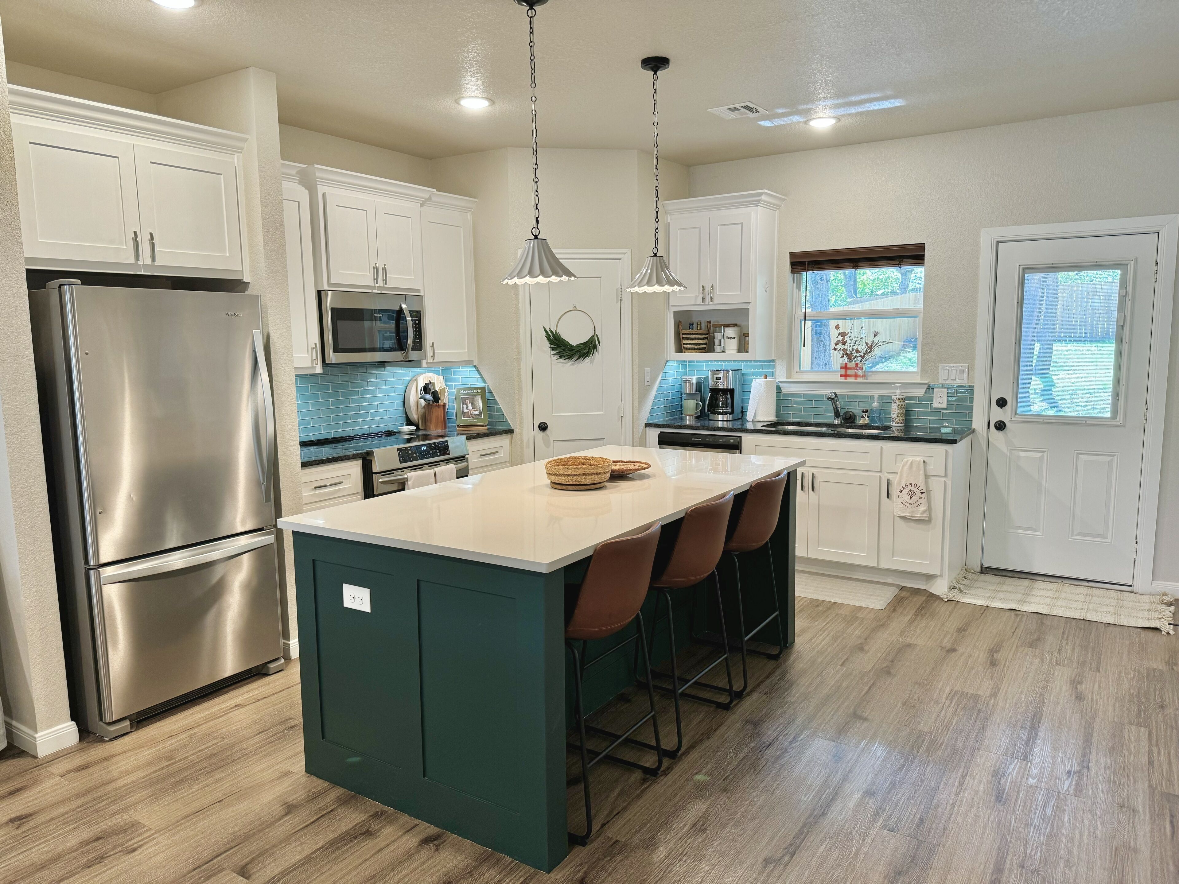 A modern kitchen with white cabinets, stainless steel appliances, a central island with bar stools, and a view to the outside.