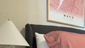 Una cama bien hecha con ropa de cama rosa y blanca, una lámpara blanca sobre una mesita de noche y un mapa enmarcado de Waco, Estados Unidos, en la pared.