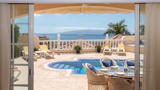 Zona de piscina con sillones de descanso y una mesa, vista al mar y una valla blanca.