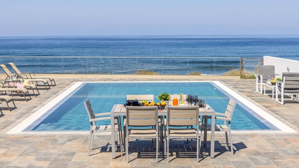 Un área de piscina con mesa y sillas, vista al océano y sillas reclinables.