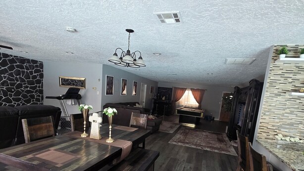 A spacious living room with a large wooden dining table, dark wooden benches, a stone accent wall, and a modern chandelier.