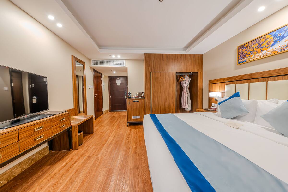A hotel room with a wooden floor, a bed with white and blue bedding, a TV, a desk with drawers, a wardrobe, and a painting on the wall.