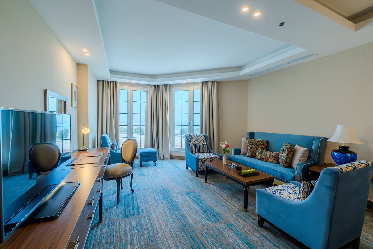 A modern hotel room with a wooden desk, a blue sofa, a blue armchair, and a large window offering a city view.