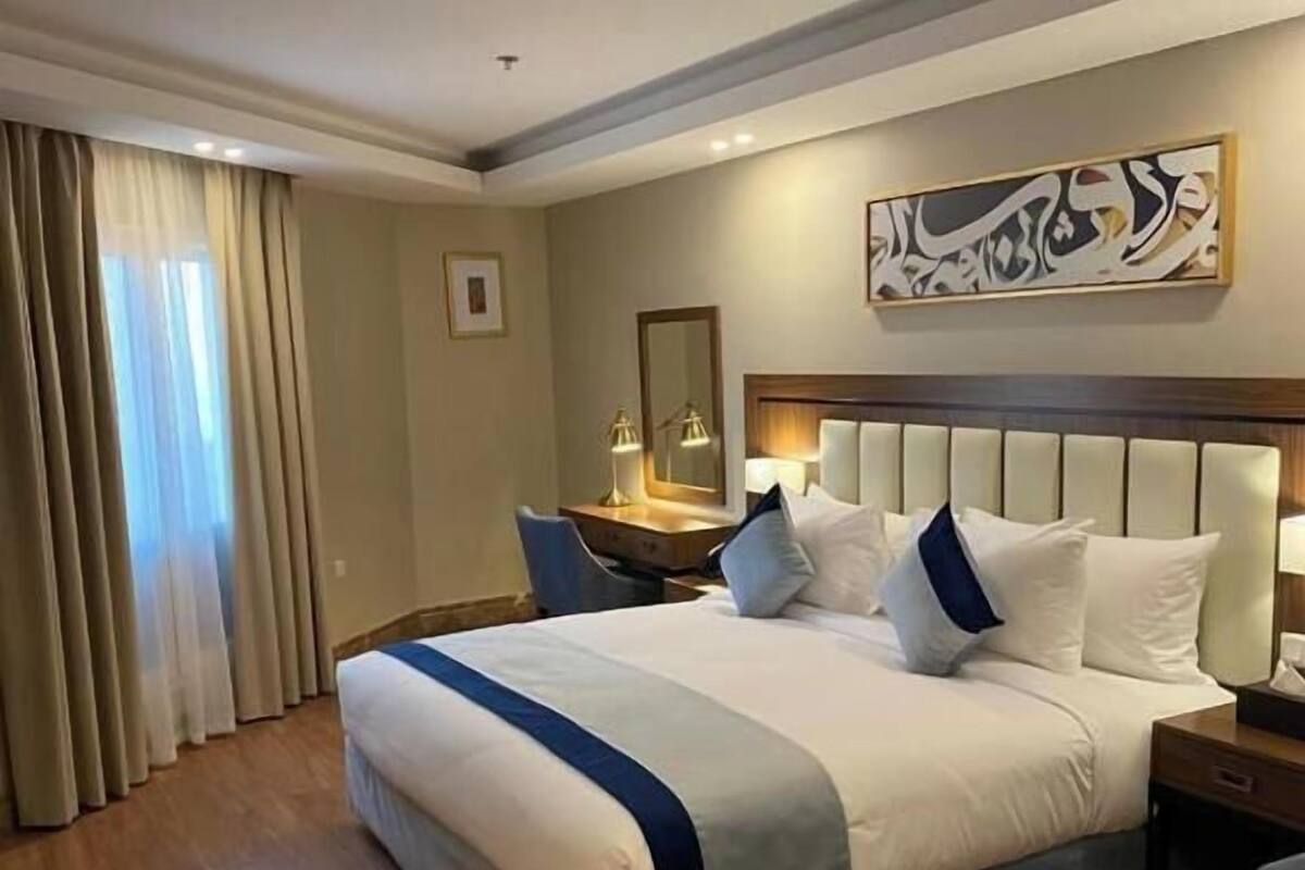 A hotel room with a large bed, a desk, a chair, a small ottoman, and a wall with Arabic calligraphy.