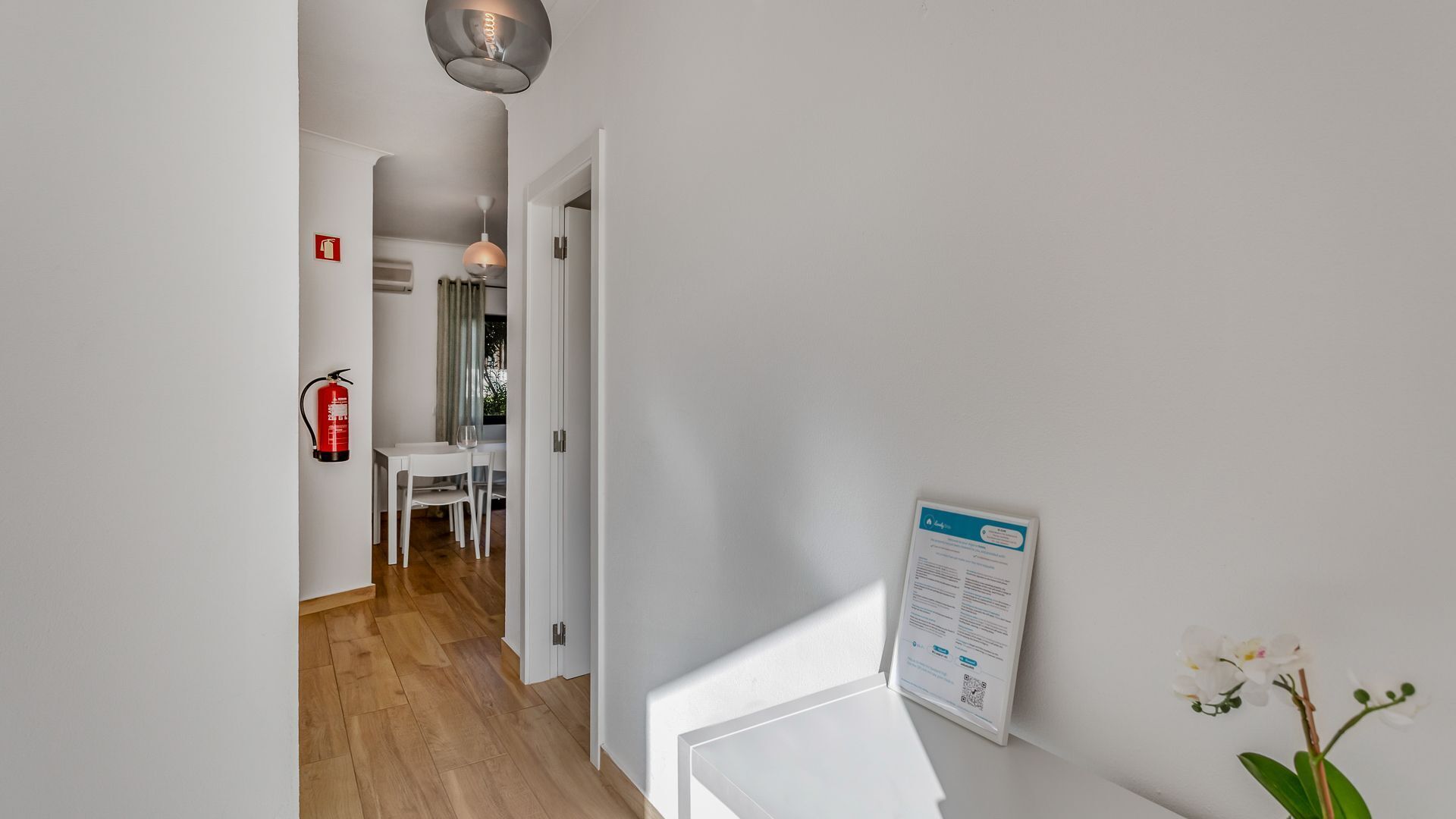 A hallway with a white wall, a fire extinguisher, a table with chairs, and a sign on the wall.