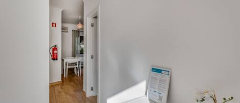 A hallway with a white wall, a fire extinguisher, a table with chairs, and a sign on the wall.