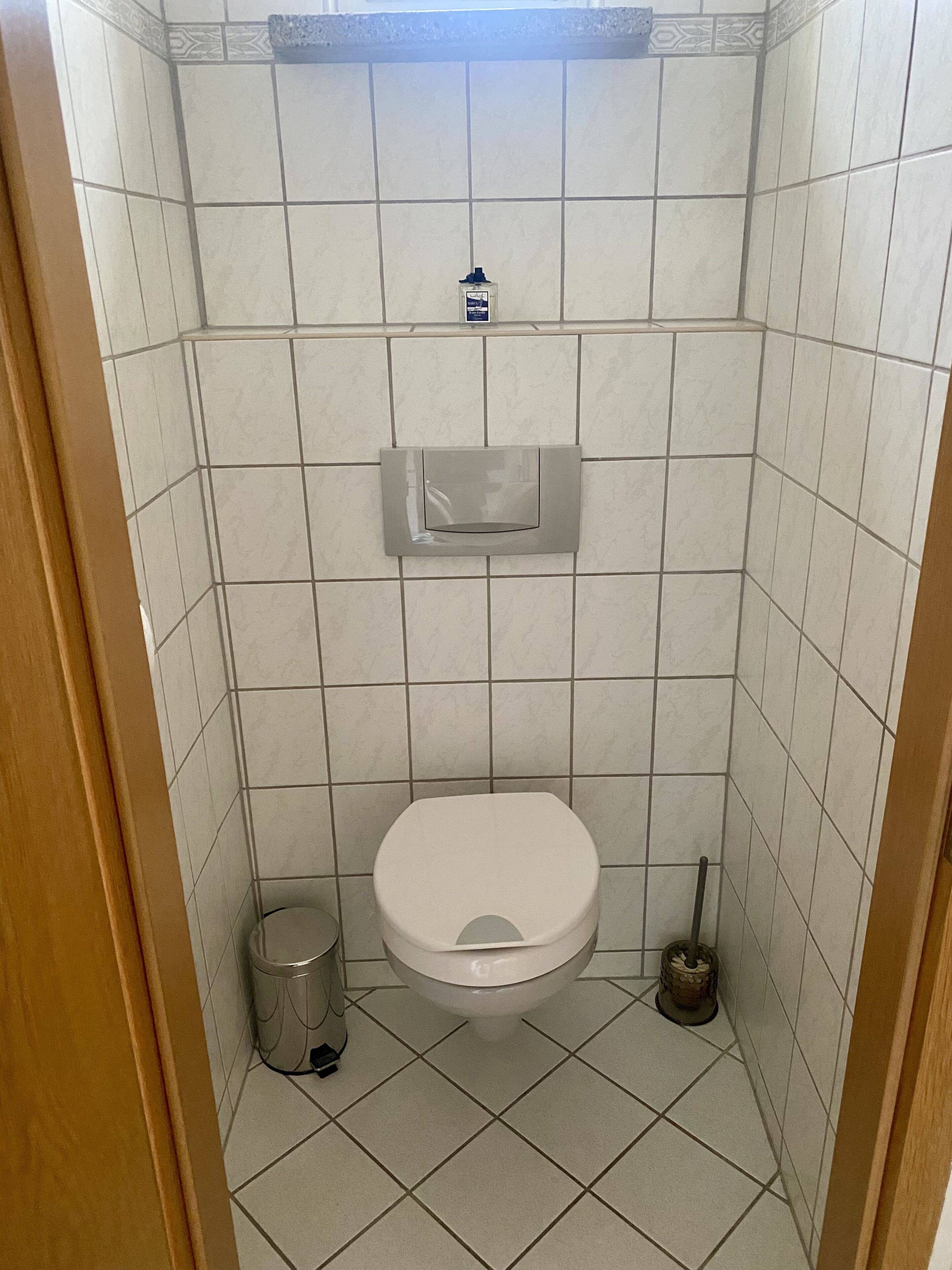 Une petite salle de bain carrelée, équipée de toilettes, d’une poubelle et d’un sèche-mains.