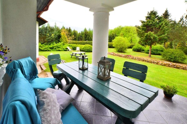A covered patio with a wooden table and chairs, a bench, and a view of a well-maintained garden.