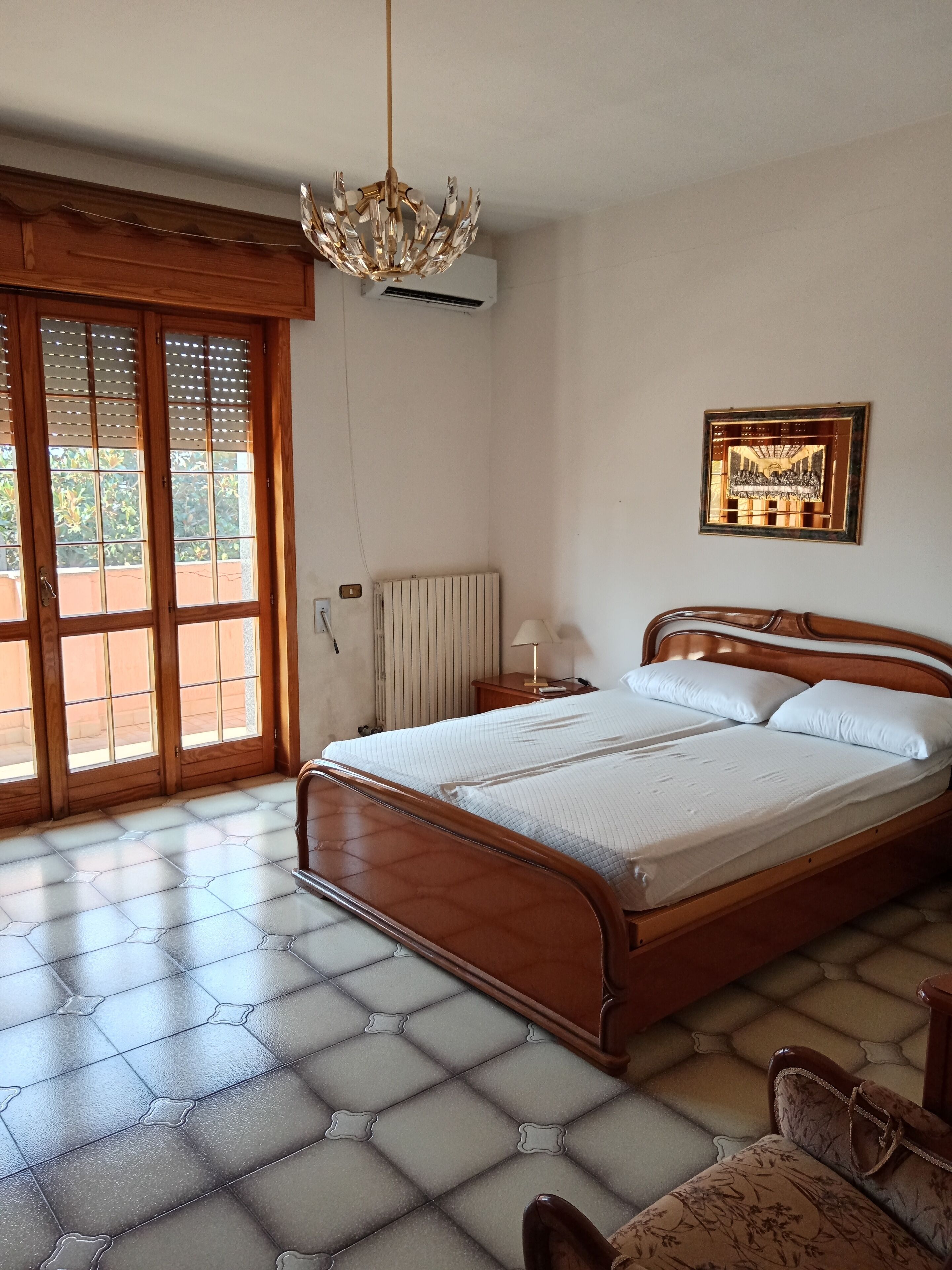 Un dormitorio con una cama de madera, un candelabro, un radiador y un amplio ventanal con persianas.