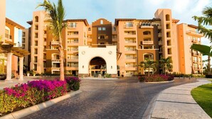Un hôtel à plusieurs étages, doté d’une entrée principale en forme d’arc central, de balcons et d’une cour pavée agrémentée de palmiers et de fleurs.
