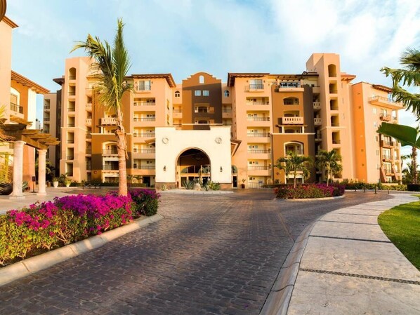A multi-story hotel with a central archway entrance, balconies, and a paved courtyard with palm trees and flowers.