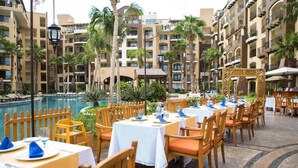 An outdoor dining area with tables and chairs, a pool, and a building with balconies in the background. - Christmas in Cabo Dec 21-29, 2024 (Cabo San Lucas)