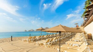 A beach with rows of sun loungers and umbrellas, a clear sky, and a view of the ocean. - Christmas in Cabo Dec 21-29, 2024 (Cabo San Lucas)