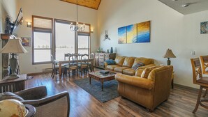 A spacious living room with a high ceiling, wooden flooring, a leather sofa, a coffee table, and a dining area with wooden chairs and a table.