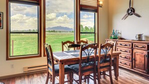 A dining area with a table and chairs, large windows with a view of a green field and mountains, and a ceiling fan.
