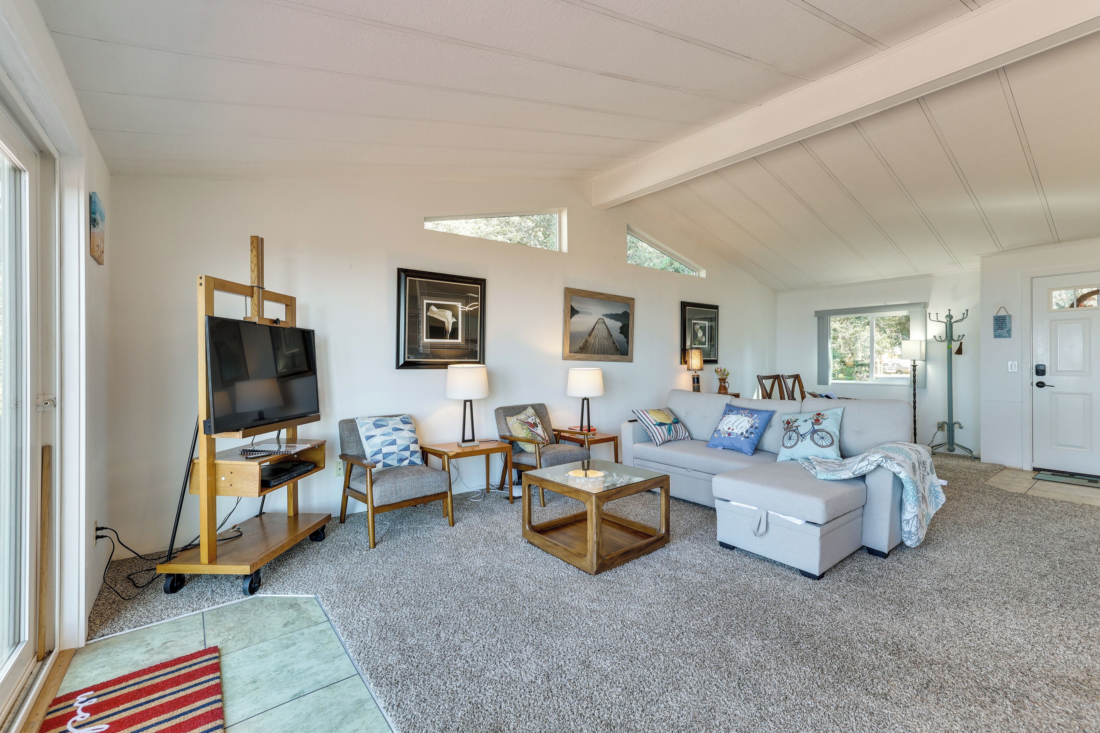 A spacious living room with a television on a wooden stand, a white sofa, two armchairs, a coffee table, and a rug.