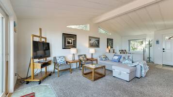A spacious living room with a television on a wooden stand, a white sofa, two armchairs, a coffee table, and a rug.