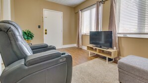 A living room with a reclining chair, a flat-screen TV on a wooden stand, a beige carpet, and a door in the background.