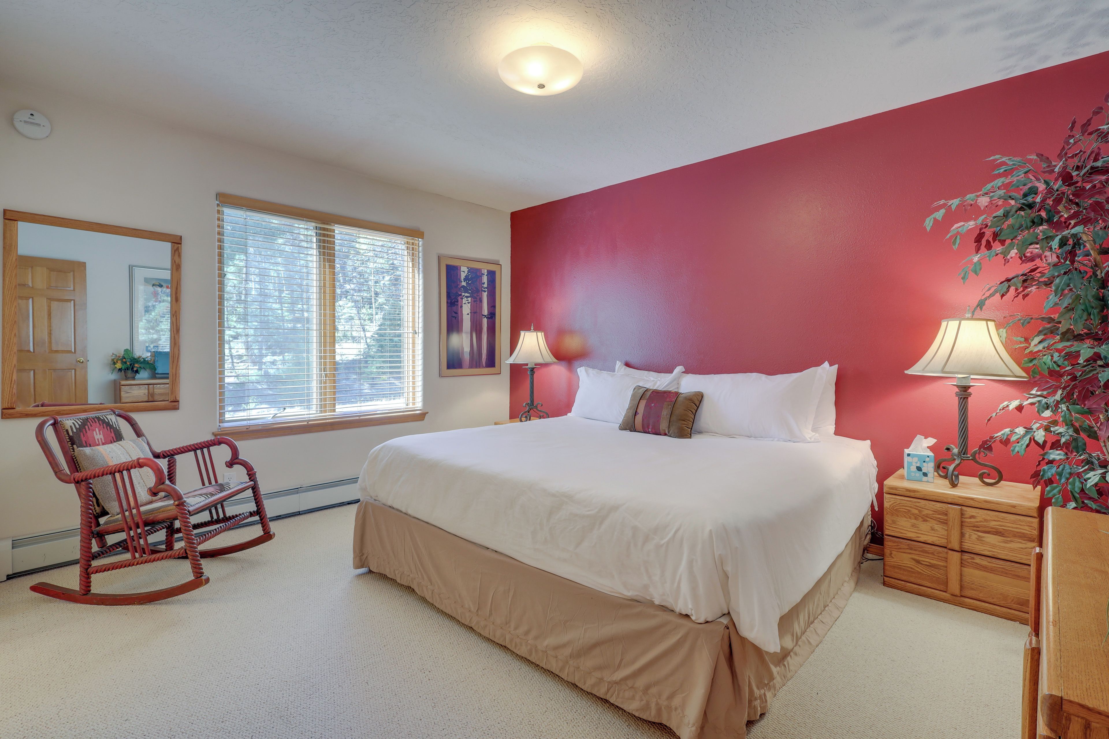 A bedroom with a large bed, a rocking chair, a nightstand with a lamp, and a window with blinds.
