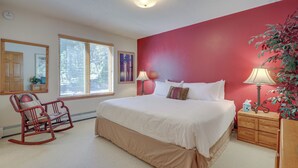 Een slaapkamer met een groot bed, een schommelstoel, een nachtkastje met een lamp en een raam met jaloezieën.