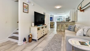 A spacious living room with a large flat-screen TV, a sofa, and a coffee table. There is a staircase leading to another level, and a kitchen area is visible in the background. - Fernandina Beach Paradise: Steps to Shore! (Fernandina Beach)