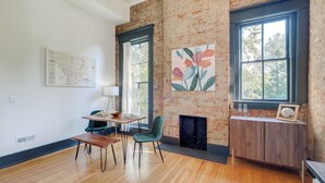 A modern living room with a brick wall, a fireplace, a dining area with a table and chairs, and a large window with a view of trees. - Chic & Historic Apartment ~ 6 Mi to Louisville! (New Albany)
