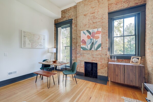 A modern living room with a brick wall, a fireplace, a dining area with a table and chairs, and a large window with a view of trees. - Chic & Historic Apartment ~ 6 Mi to Louisville! (New Albany)