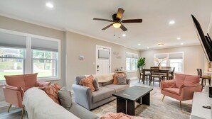 A modern living room with a grey sofa, a white sofa, and a pink armchair. There is a ceiling fan, a dining table with chairs, and a window with blinds. - Updated Atlanta Home ~ 5 Mi to Downtown! (Atlanta)
