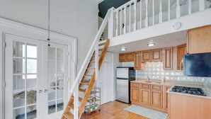 Un'ampia cucina con mobili in legno, piano cottura integrato e frigorifero. Una scala in legno conduce ad una zona mansardata con balcone.