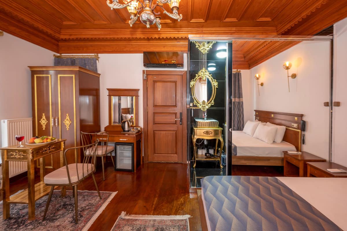 A hotel room with a wooden ceiling, a bed, a desk with a chair, a wardrobe, and a mirror.