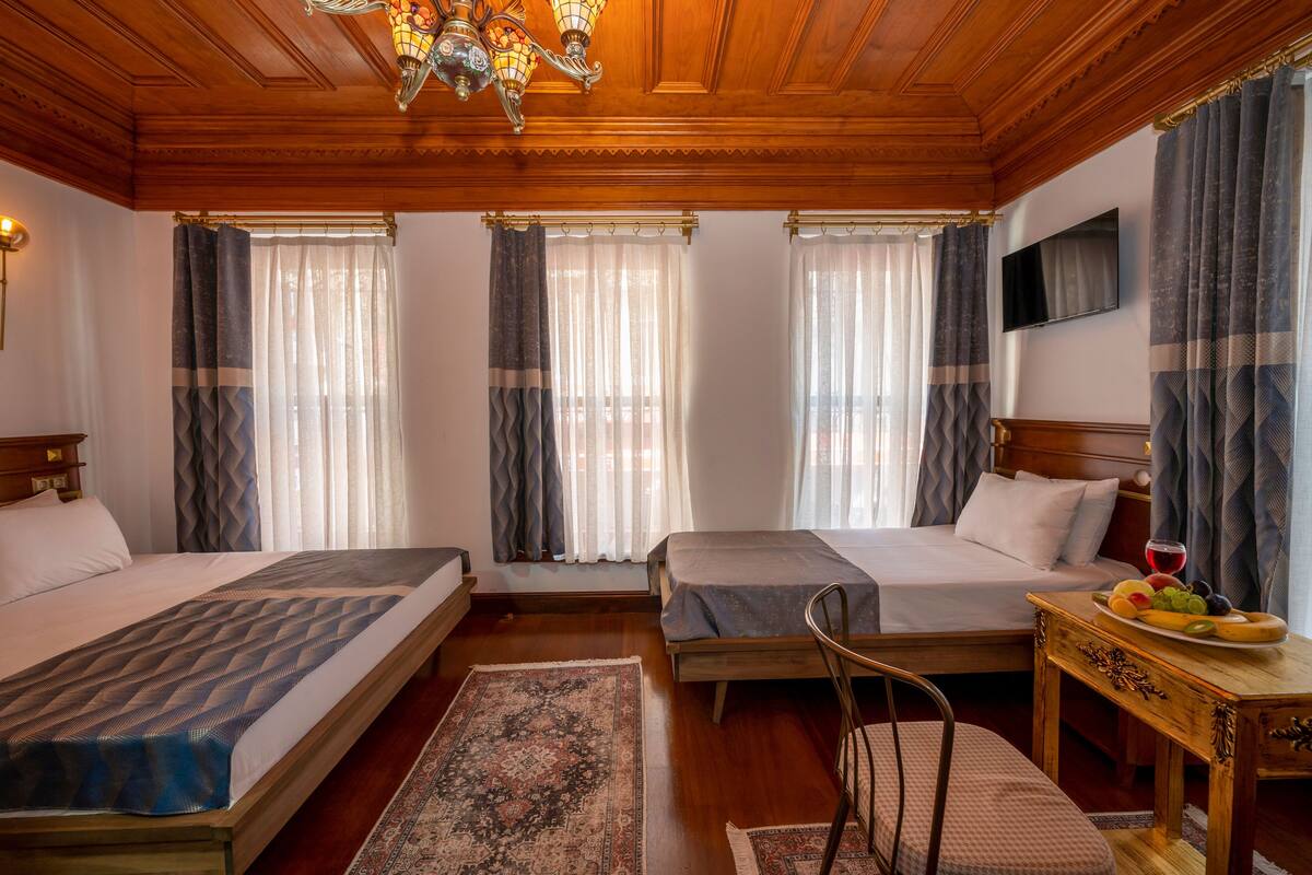 A hotel room with two beds, a wooden ceiling, a chandelier, a flat-screen TV, and a small table with fruit.