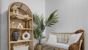 A wicker sofa with a brown throw blanket, a wicker side table with decorative items, and a large potted plant.