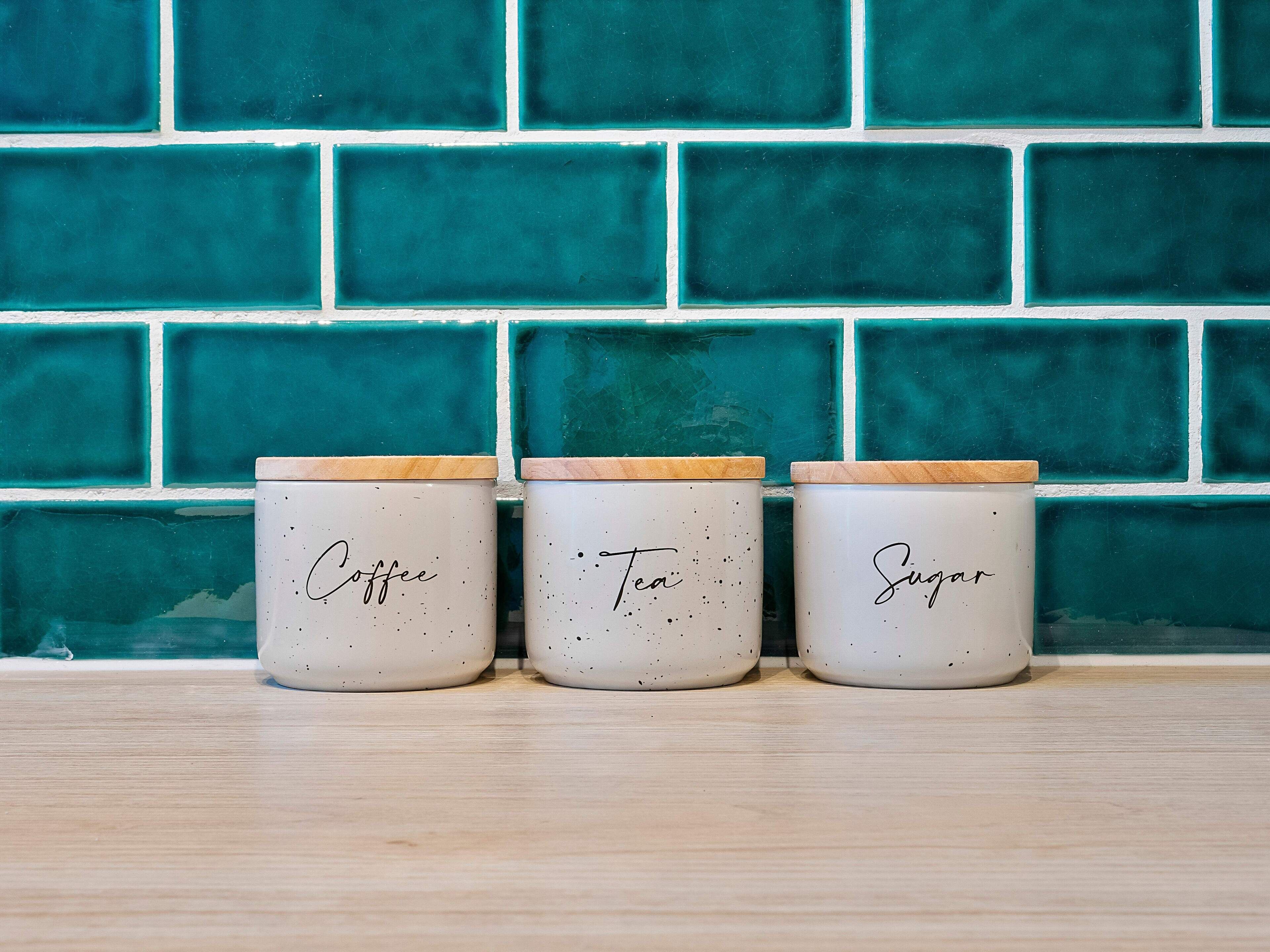 Three ceramic containers with wooden lids labeled "Coffee," "Tea," and "Sugar" on a wooden surface against a teal tiled wall.