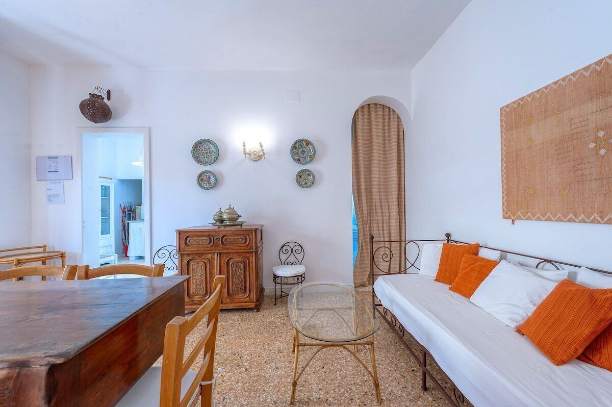 A room with a wooden dining table, a sofa with orange pillows, a glass table, and a cabinet with decorative plates on the wall.
