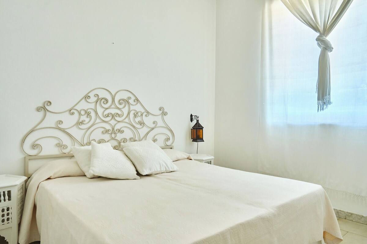 A neatly made bed with a white headboard and pillows, a bedside table with a lamp, and a window with a curtain.