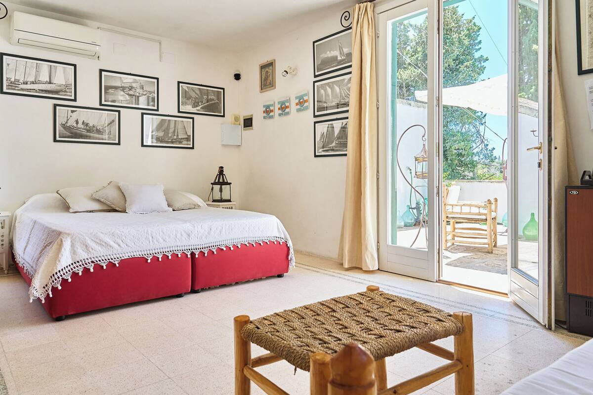 A bedroom with a bed, a red bench, and a balcony with a view of the outdoors.