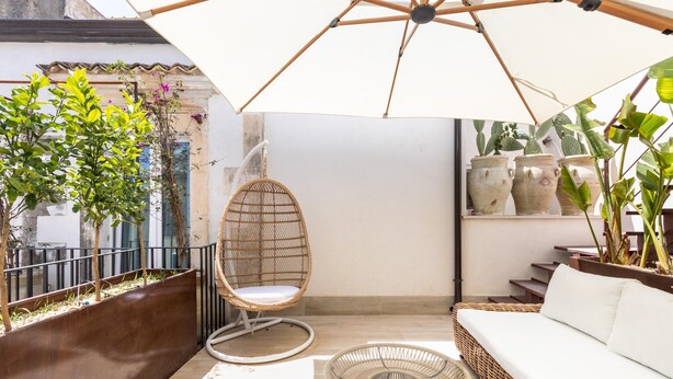 A sunlit patio with a wicker sofa, a hanging chair, and potted plants. - Bowie's Rooms (Noto)