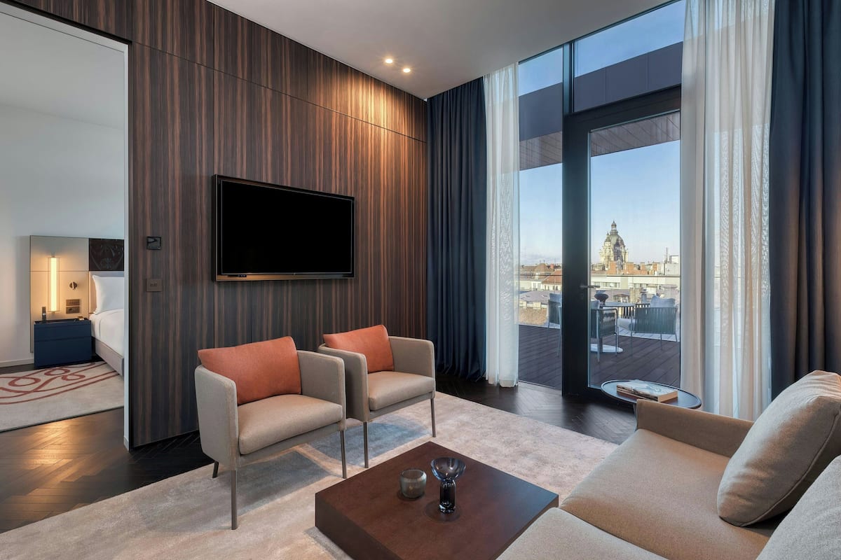 A modern living room with a large TV, two armchairs, a sofa, and a view of a cityscape through large windows.