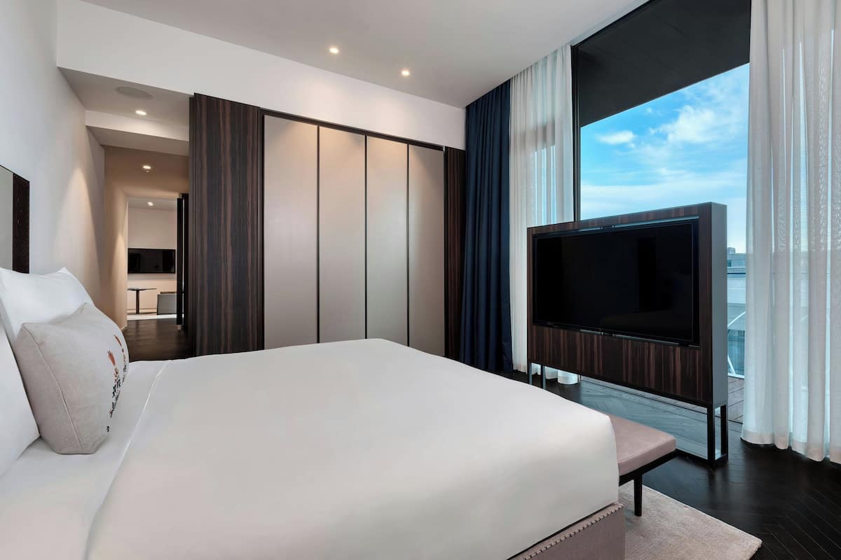 A modern hotel room with a large bed, a flat-screen TV mounted on the wall, and a view of the city through large windows.