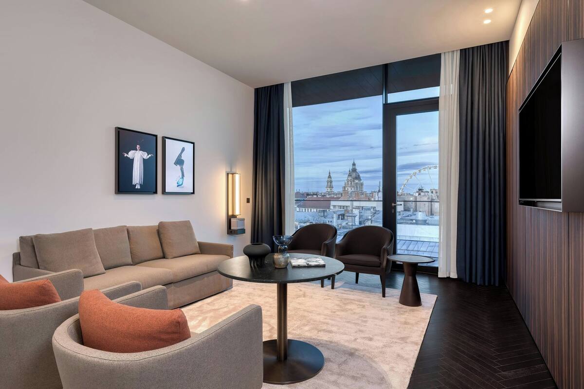 A modern hotel room with a sofa, armchairs, a round table, and a large window offering a city view.